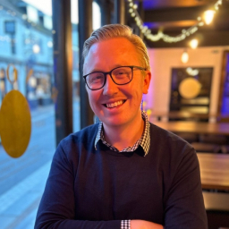 A smiling man with blond hair and glasses wears a navy sweater and checkered shirt, indoors.