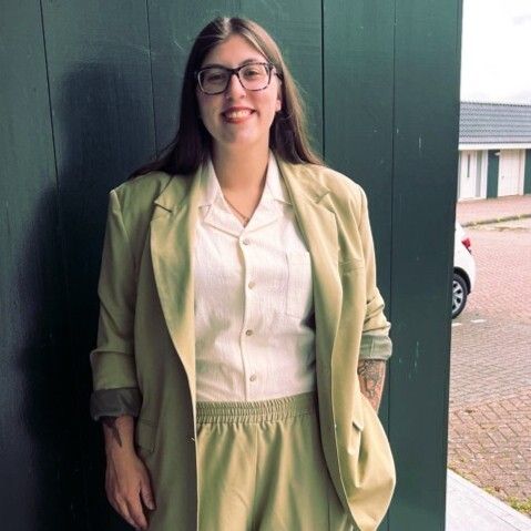 A smiling woman in glasses and a light green suit stands by a dark green wall.