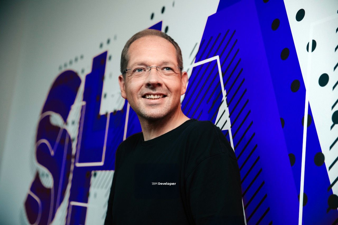 A man wearing an IBM Developer shirt and glasses smiles in front of an abstract blue and white background.