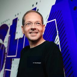 A man wearing an IBM Developer shirt and glasses smiles in front of an abstract blue and white background.
