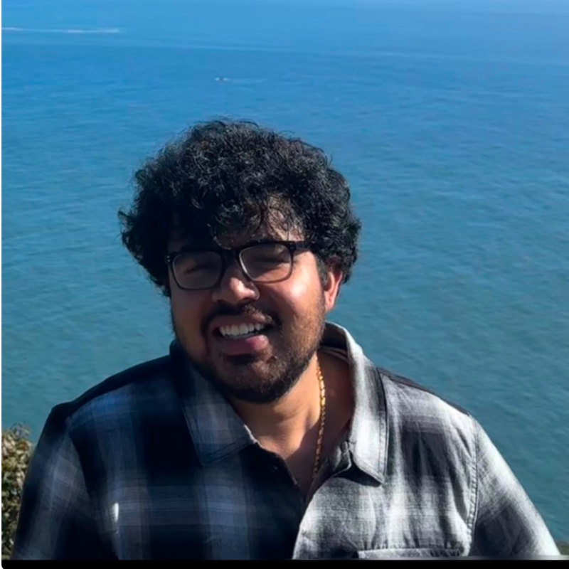 A smiling man with curly dark hair and glasses in a plaid shirt with the ocean behind him.