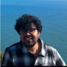A smiling man with curly dark hair and glasses in a plaid shirt with the ocean behind him.