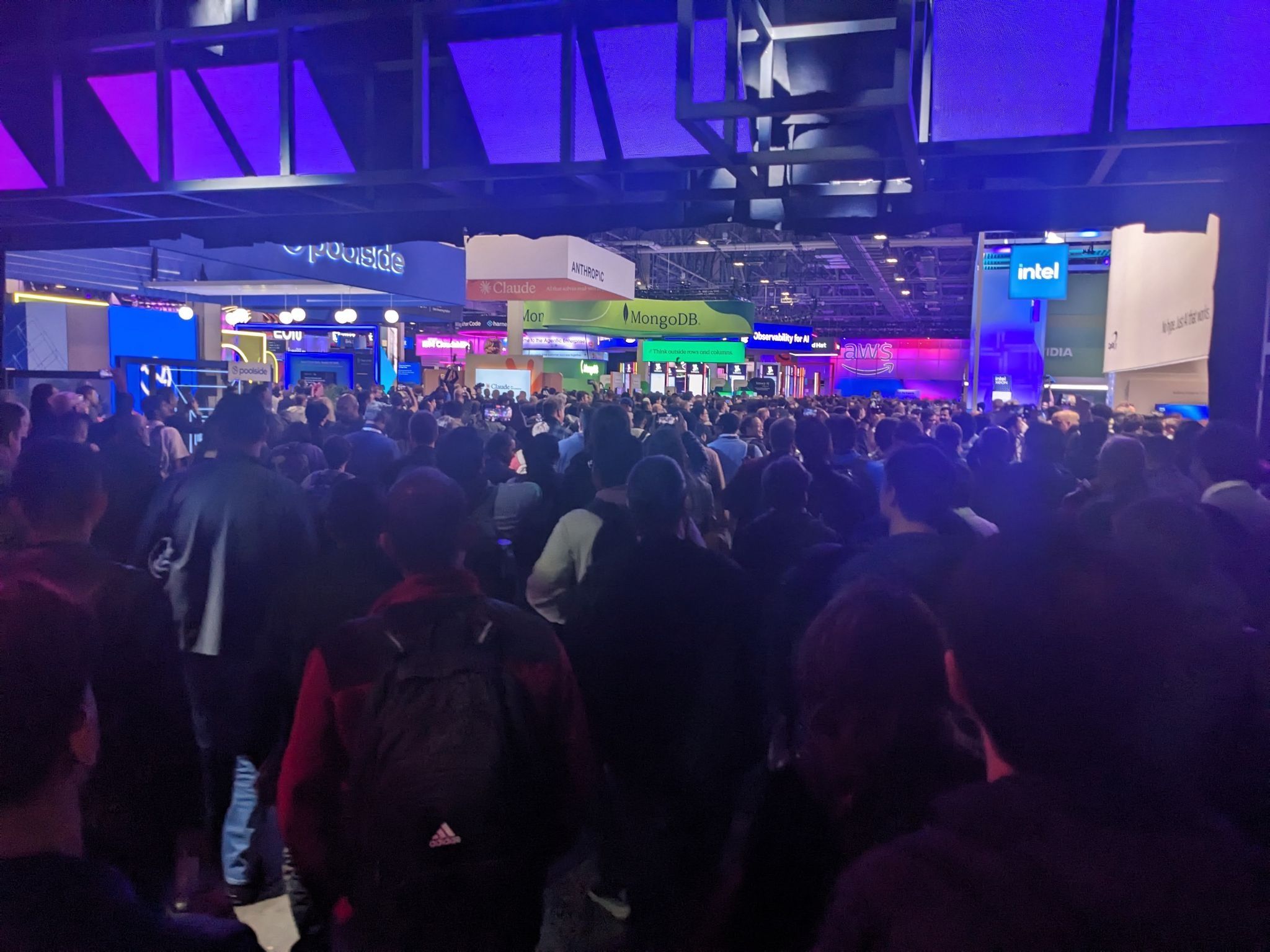 A crowd of people walking toward a hall filled with company booths. The booths display brand names such as "Anthropic," "Claude," "MongoDB," "Iogear Code Share," "AWS," and others. The scene reflects a bustling setting at the AWS re:Invent 2025 convention, with themes of artificial intelligence, technology, and innovation.
