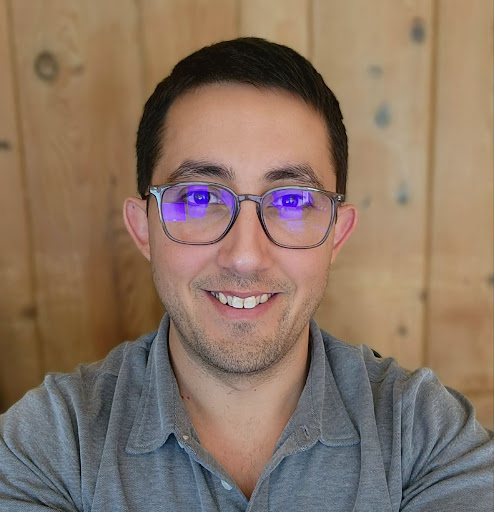 A smiling man with dark hair wears glasses with purple reflections and a gray polo shirt against a wooden background.