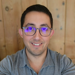 A smiling man with dark hair wears glasses with purple reflections and a gray polo shirt against a wooden background.